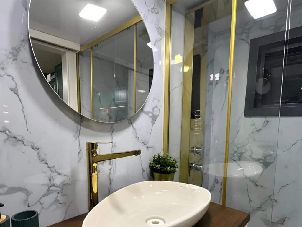 Luxurious tiny home bathroom with marble-effect wall tiles, a sleek oval vessel sink, a large circular mirror, and modern gold-finish faucet and shower frame.