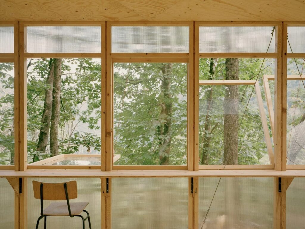 Interior view of Casetta Tessino looking out through large wooden-framed windows at the vibrant green forest, with a minimalist desk and chair.