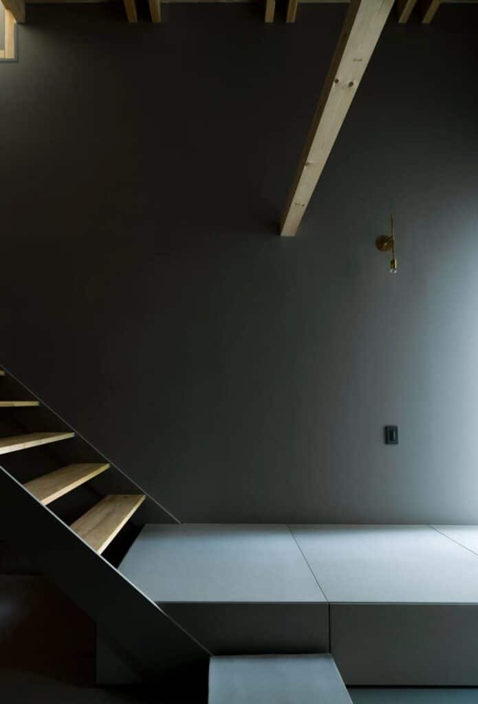 Minimalist wooden stairs against a dark grey wall in Fukasawa House, highlighting shadow and light.