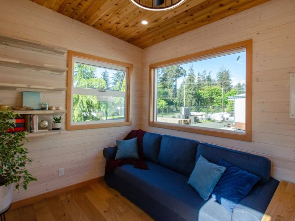 Interior living area of Juniper tiny house with a large blue sofa, wooden walls, and expansive windows.