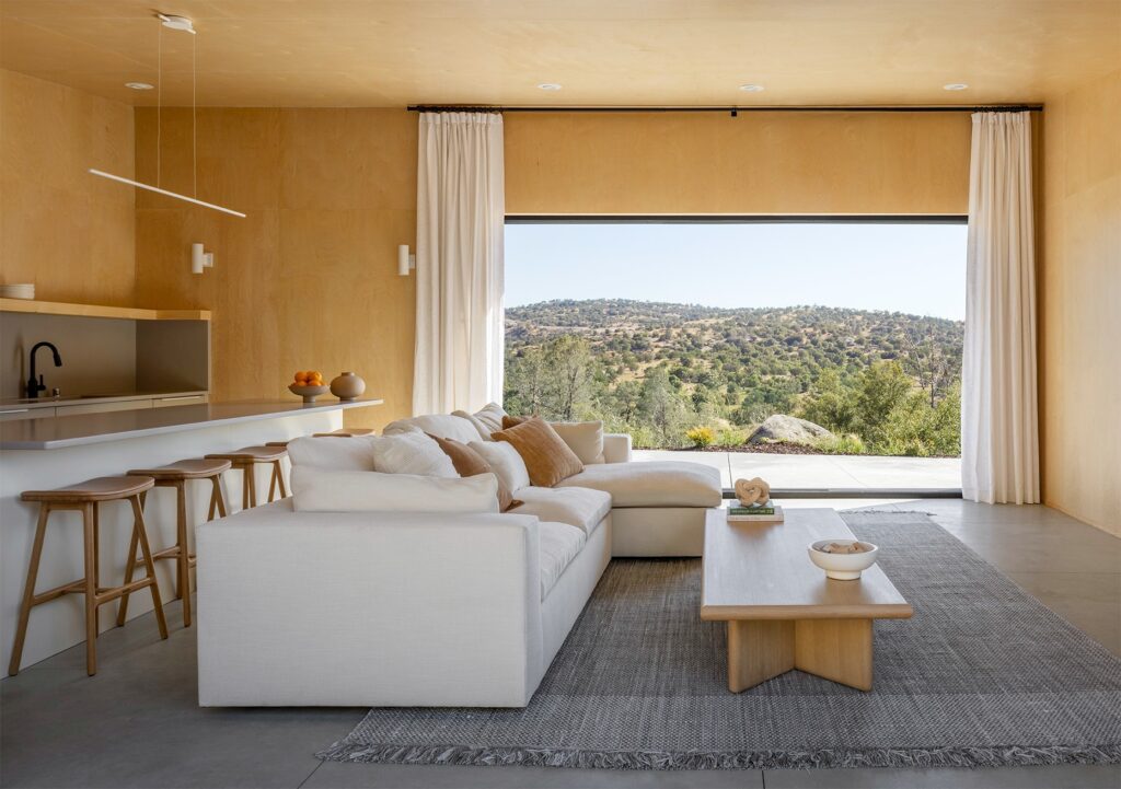 Living room interior of Casa Roca featuring a white sectional sofa, plywood walls, and a large panoramic window framing the mountain horizon.