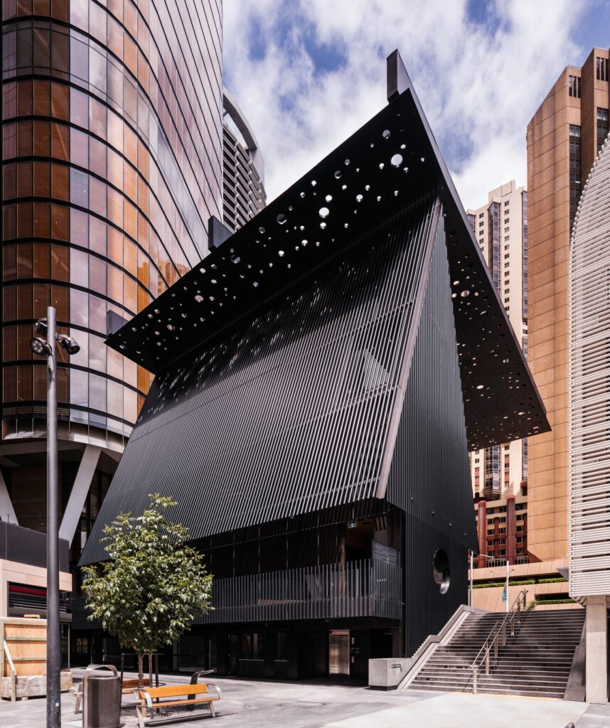 Close-up exterior view of the George Street Community Building, showcasing its sharp black facade, vertical louvers, and a portion of the perforated canopy against a backdrop of modern skyscrapers.