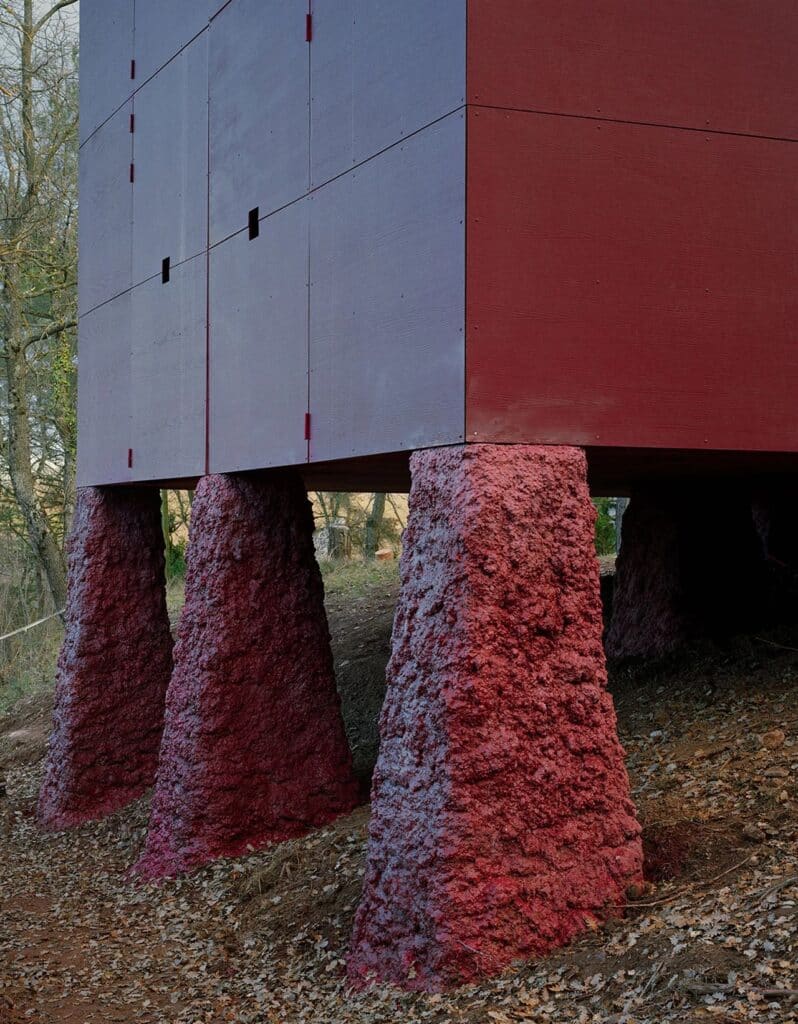View of the red Prefab Changeover house through the branches of a large, bare tree in winter, on a wooded slope.