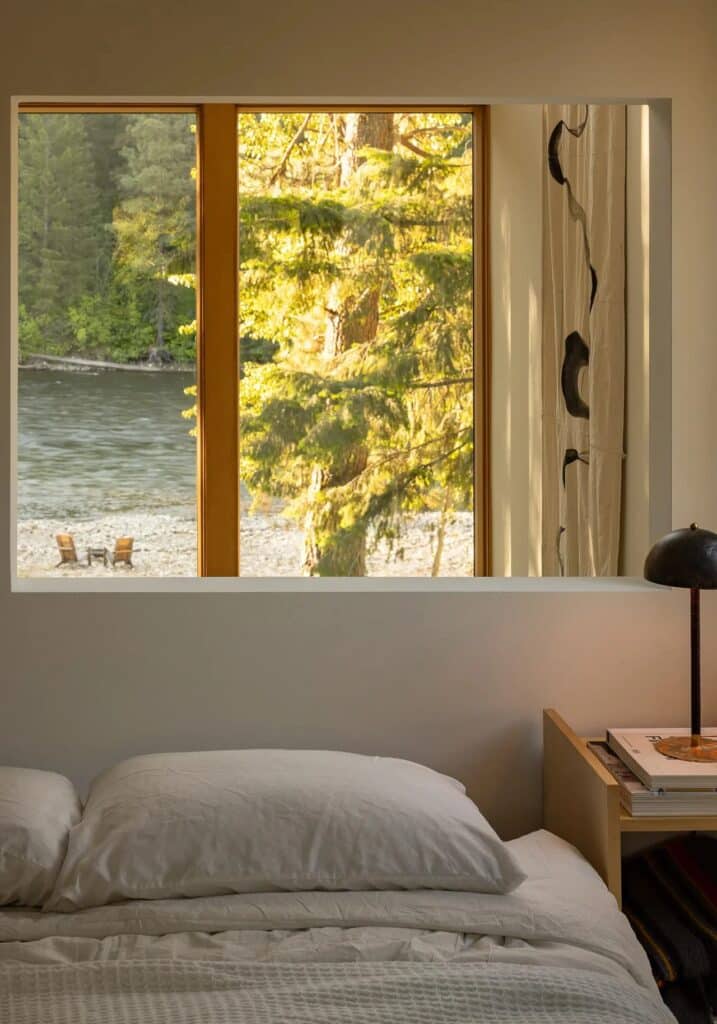 Bedroom interior view with a large window framing a sunlit pine tree and the Wenatchee River and two chairs on the distant bank.