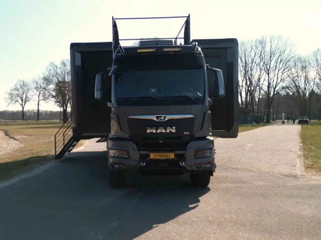 Frontal view of the dark grey MAN truck carrying the Shiver x Expandable unit, showing its wide stance and deployed side modules.