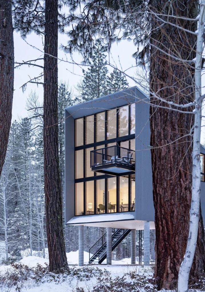 Wide exterior view of the Wenatchee River Cabin in winter, partially obscured by a line of tall, snow-dusted pine trees, overlooking the snowy riverbank.