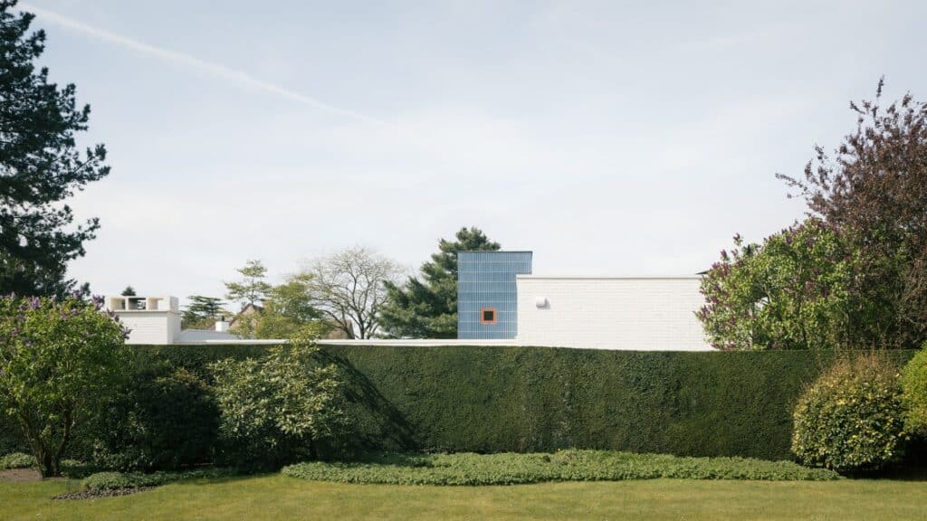 View of Bungalow Daniel from a distance behind a tall green hedge, showing the top of the blue tower and white brick facade.