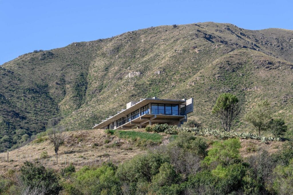 A brutalist concrete house perched on a hillside in Punilla Valley, showing the relationship between the structure and the vast mountain landscape.