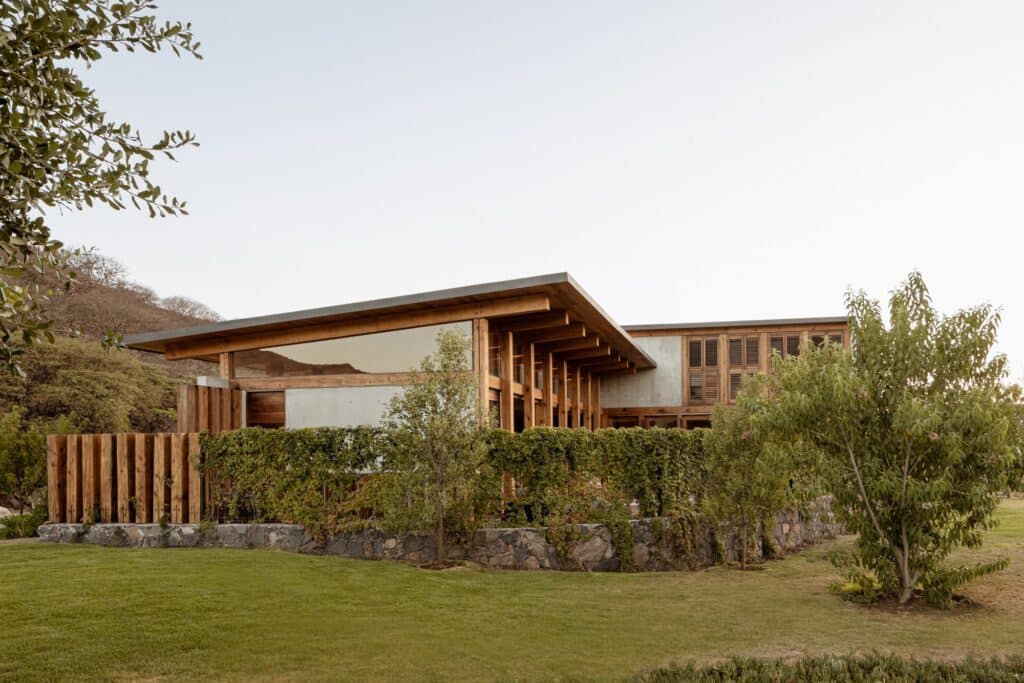 Side view of Queretaro House with a stone and green hedge fence, showcasing the flat roof and timber columns.