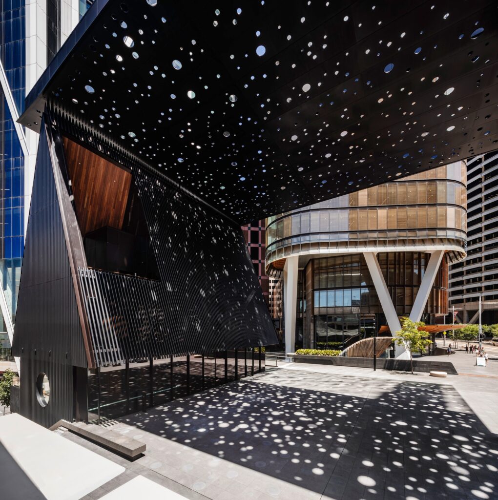 View under the George Street Plaza's perforated black canopy, showing the dappled light patterns cast onto the ground and the open side of the community building.