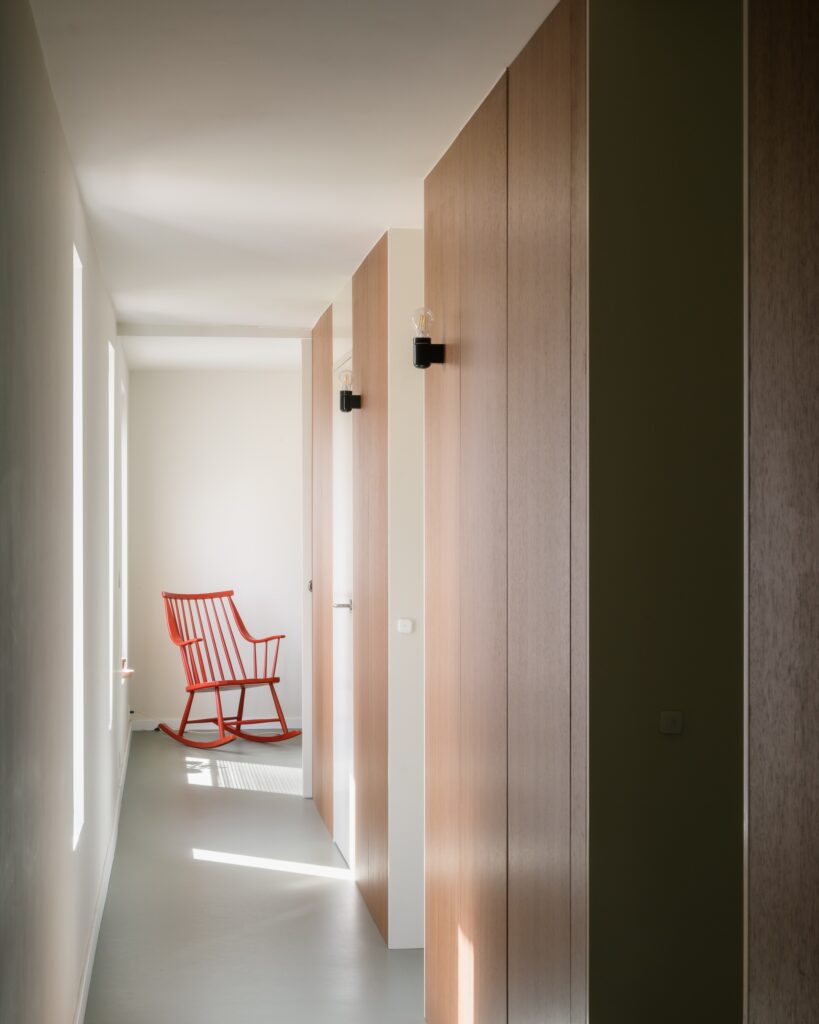 Minimalist white corridor with wooden doors and a bright red rocking chair at the end of the hallway.