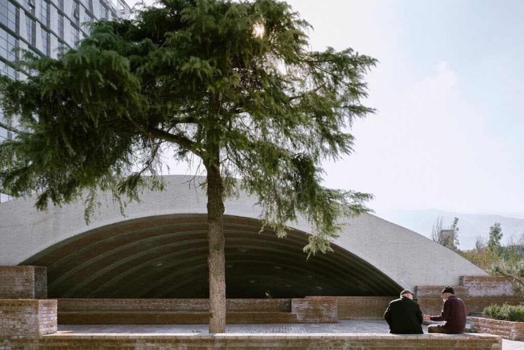 Large concrete arched opening with a shaded courtyard area and people sitting on brick ledges.