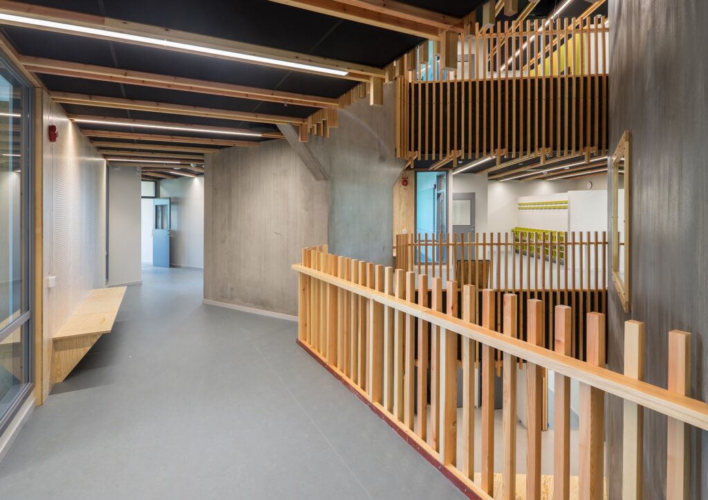 Interior corridor of &Auml;ngsdal School showing concrete walls, wooden vertical slats for railings, and modern acoustic ceiling treatments.
