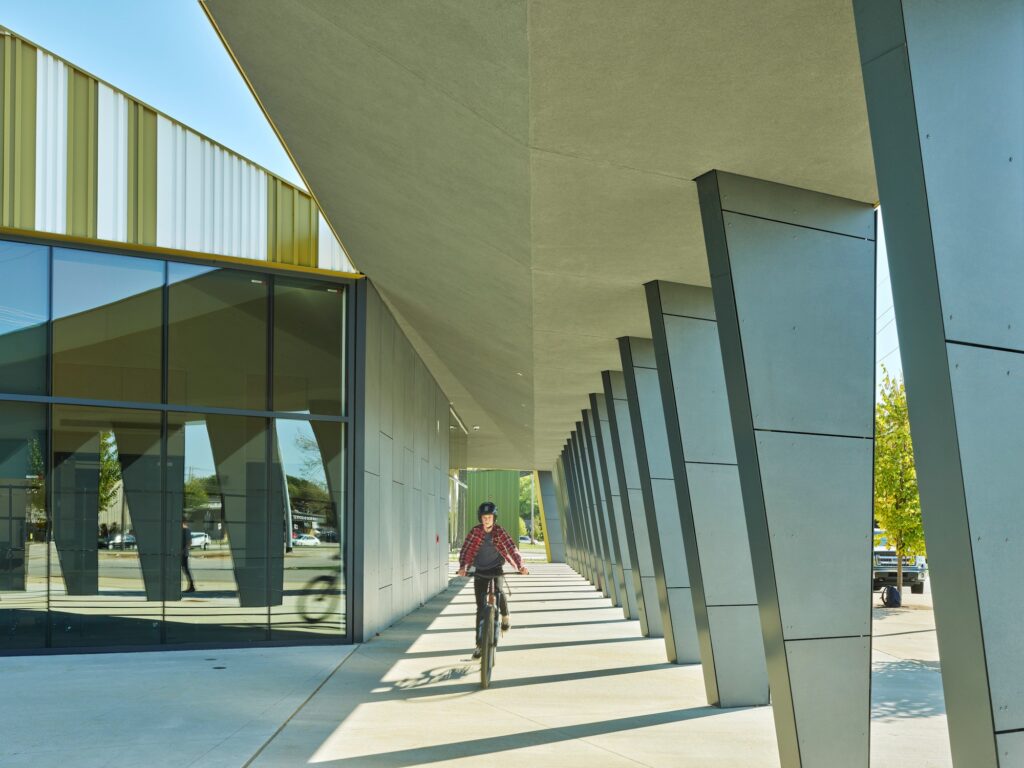 A student on a bicycle riding through a long outdoor walkway at Thaden School with dramatic tilted gray pillars and glass windows.