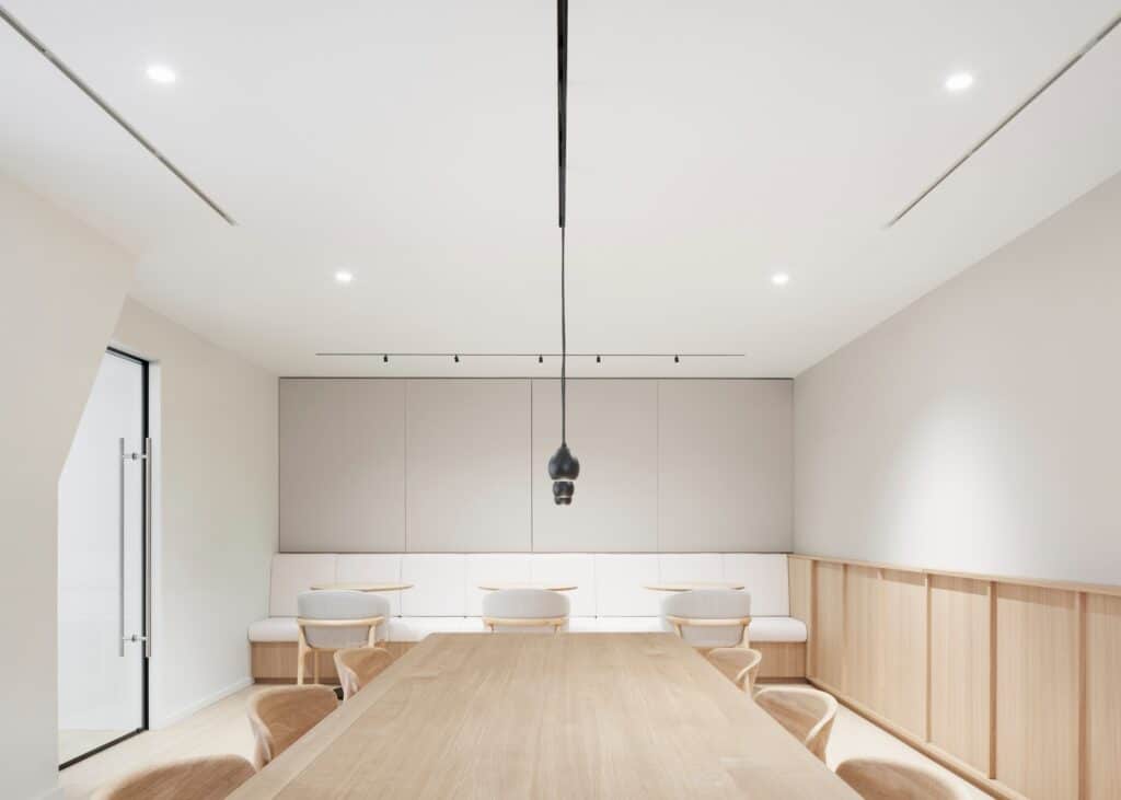 A long timber meeting table in a bright minimalist room with built-in seating and modern pendant lights.