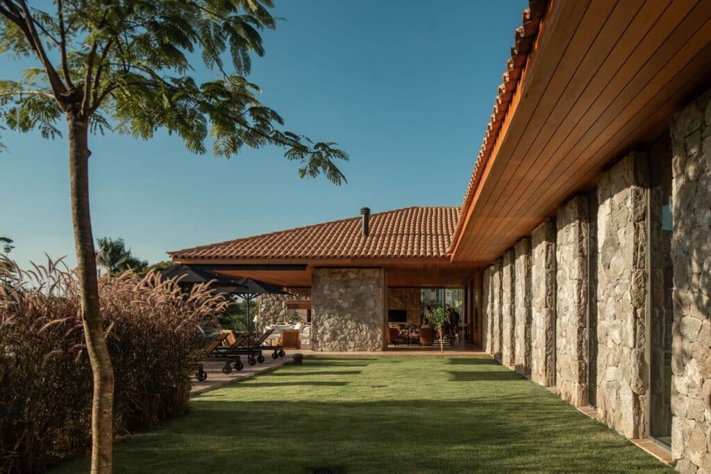 Side view of Stone House in S&atilde;o Paulo featuring rustic stone walls, a long wooden eave, and a manicured green lawn under a clear blue sky.