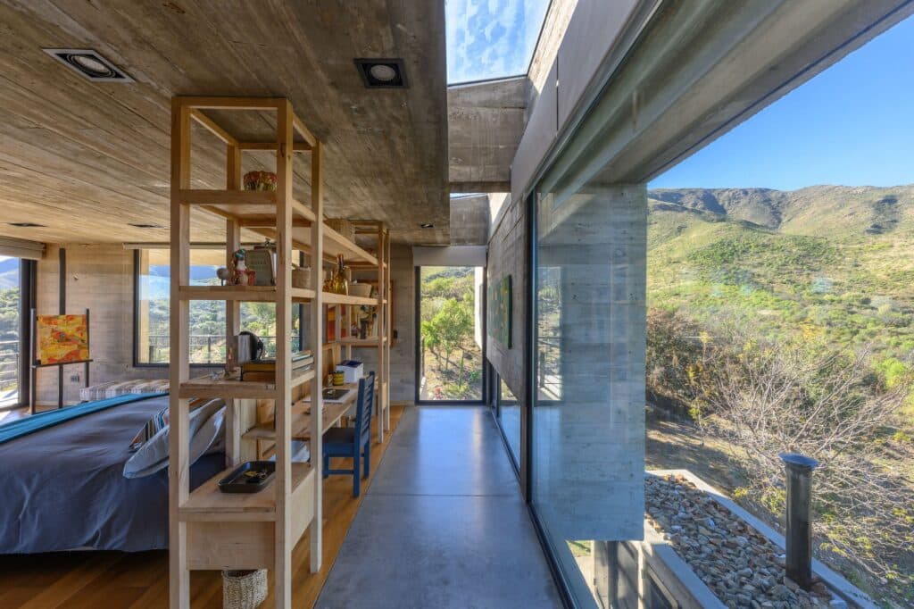Interior view of a master bedroom in Casa El Drag&oacute;n featuring exposed concrete walls, a wooden shelving unit, and a large window overlooking the mountains.