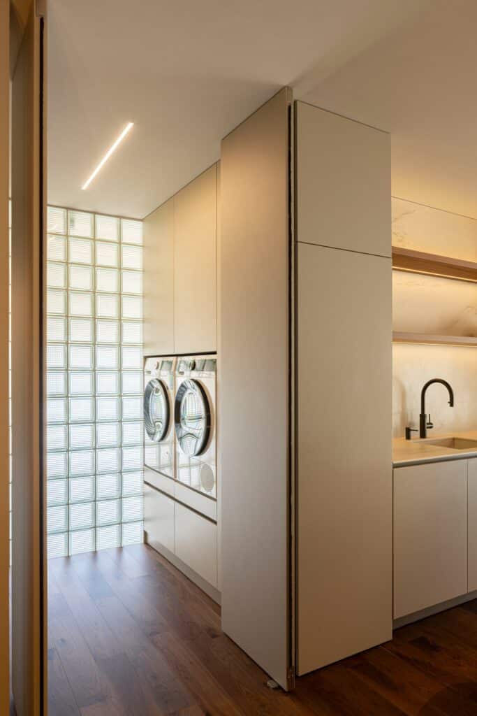 Integrated laundry area in Casa do Engenho with stacked washer and dryer, glass block wall, and minimalist white cabinetry.