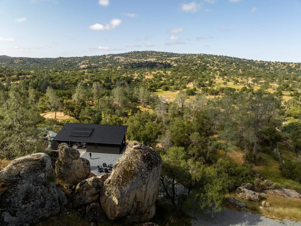 High-angle aerial view of Casa Roca, a black modern house nestled within a lush green mountainous valley with scattered rock formations.