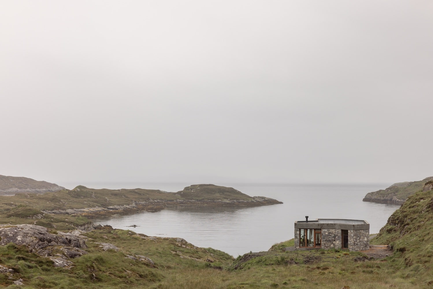 A secluded, low-lying modern stone cabin with a flat roof nestled into a grassy hillside overlooking a misty, rocky Scottish coastal inlet