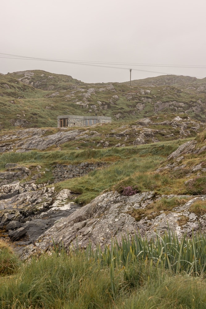A low-profile, stone-clad house, Caochan na Creige, blends into the rocky, grassy, and misty coastal landscape of the Isle of Skye, Scotland.