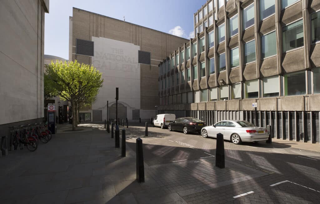 A street view showing a brick extension of the National Gallery, with the name painted on a large white section, adjacent to a modern concrete office building. A small green tree stands to the left, and two cars and a van are parked along the curb.