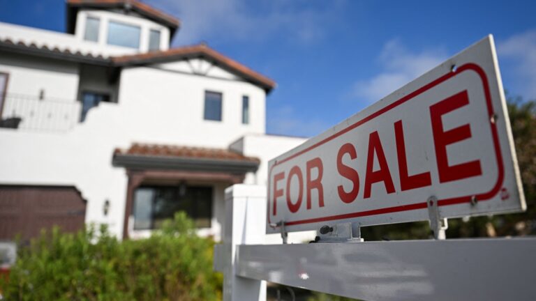 A close-up, selective focus shot of a white and red "FOR SALE" sign in the foreground, with a large, two-story white modern house blurred in the background under a blue sky.