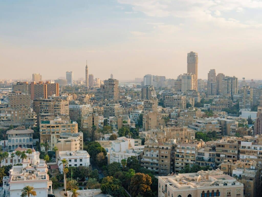 An elevated, wide-angle view of the Cairo cityscape during golden hour, featuring a dense mix of residential buildings, greenery, and the iconic Cairo Tower standing prominently in the background under a soft, hazy sky.