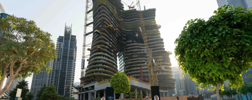 A massive twin-tower skyscraper under construction with a large archway connecting the two curved structures, surrounded by other high-rise buildings and prominent trees.