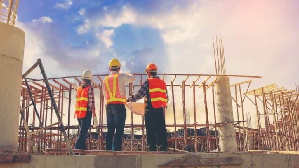 Three construction professionals wearing hard hats and safety vests standing on a site and looking at building plans against a bright sky.