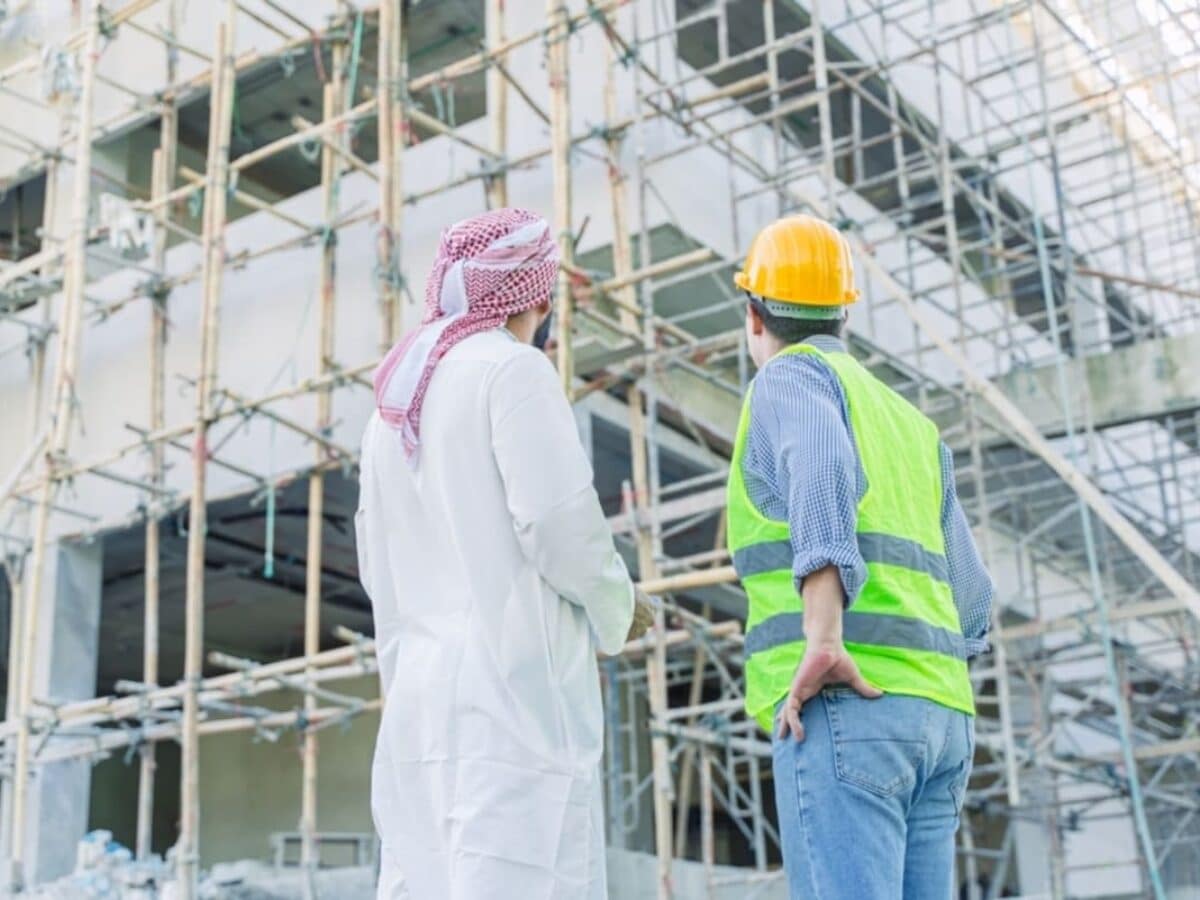 A man in traditional Saudi attire and an engineer in a safety vest and yellow hard hat looking at a building under construction with extensive scaffolding.