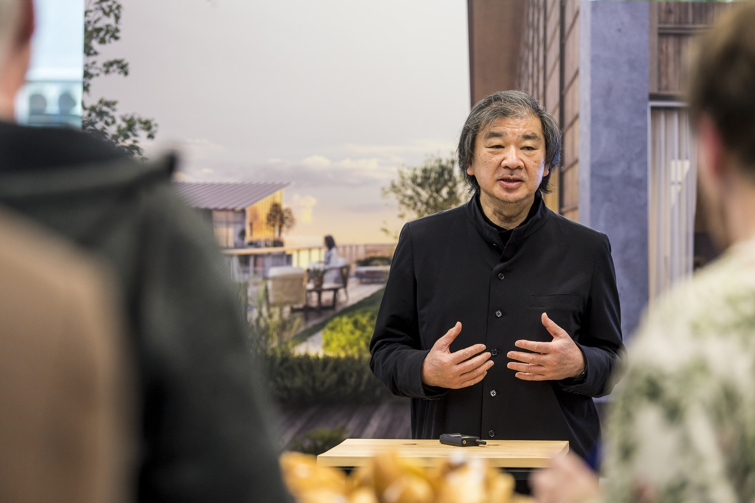 شيغيرو بان يتحدث في فعالية مع وجود مجسمات ومشاريع معمارية في الخلفية.