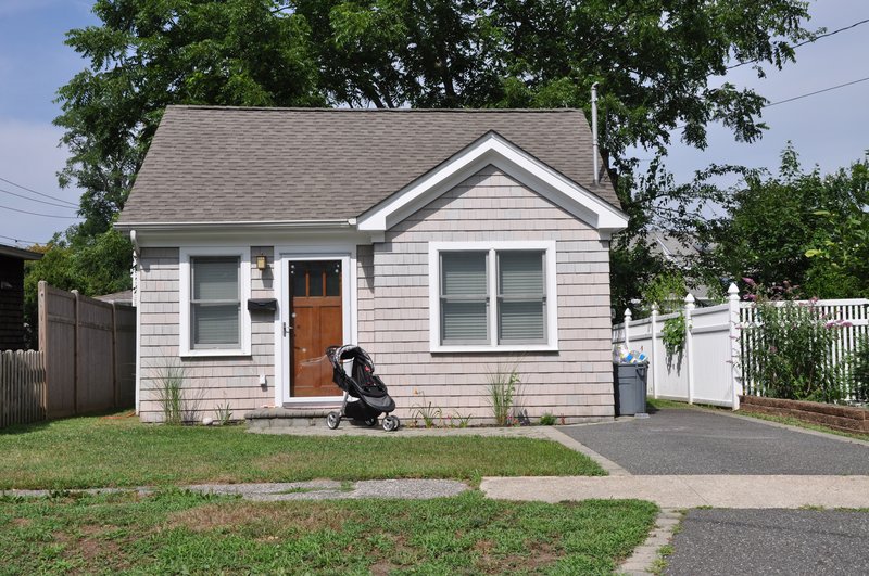 A small, single-story, grey shingle-sided house with a peaked roof and a brown front door sits between a wooden fence on the left and a white picket fence on the right. A black stroller is parked near the front door.