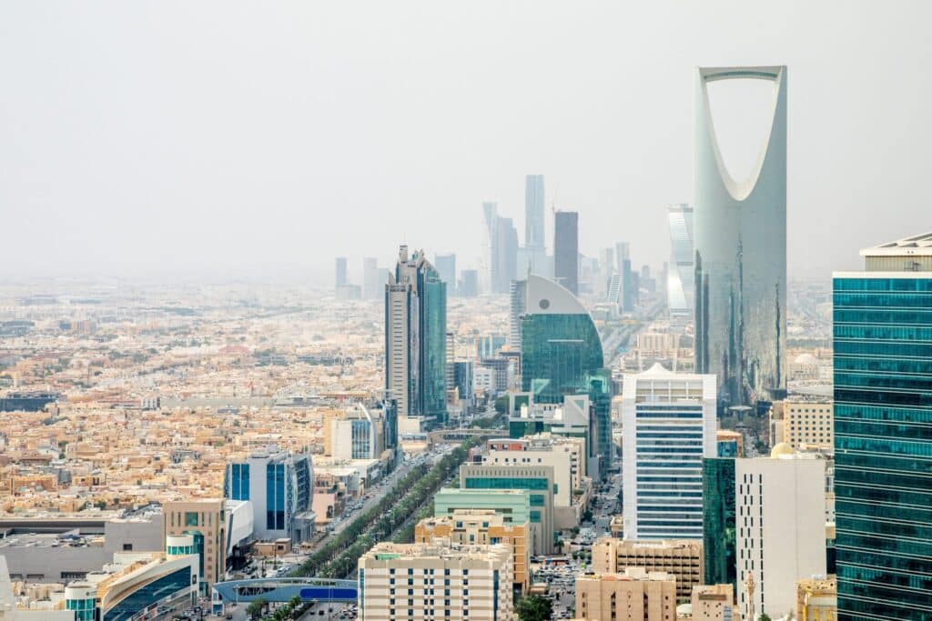 High-angle aerial view of the Riyadh skyline featuring the Kingdom Centre and Al Faisaliyah Center under a bright, hazy sky.