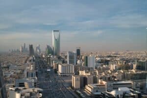 An aerial view of the Riyadh skyline featuring the prominent Kingdom Centre skyscraper and Al Faisaliyah Centre, surrounded by a sprawling cityscape and a busy highway.