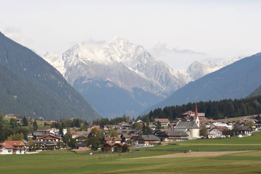 A peaceful alpine village with traditional houses and a tall church steeple set in a green valley, surrounded by forested hills and snow-covered mountain peaks in the background.