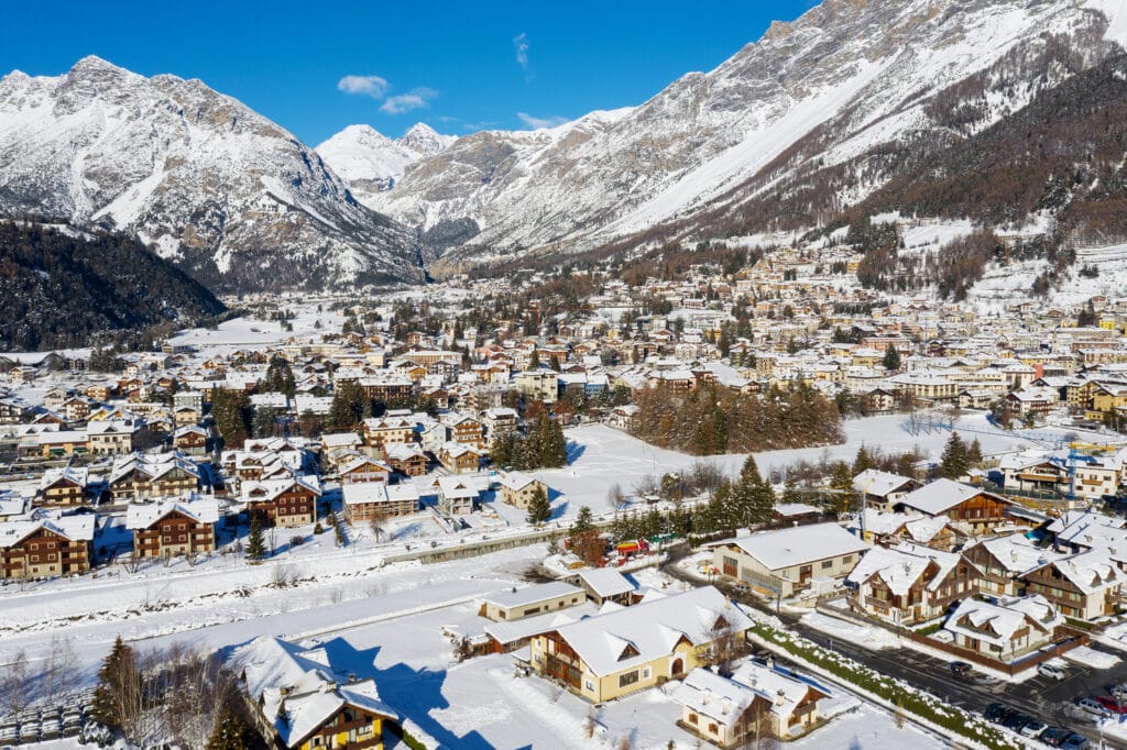 A snowy alpine town surrounded by tall, rugged mountains under a clear blue sky, with houses and buildings covered in fresh snow.