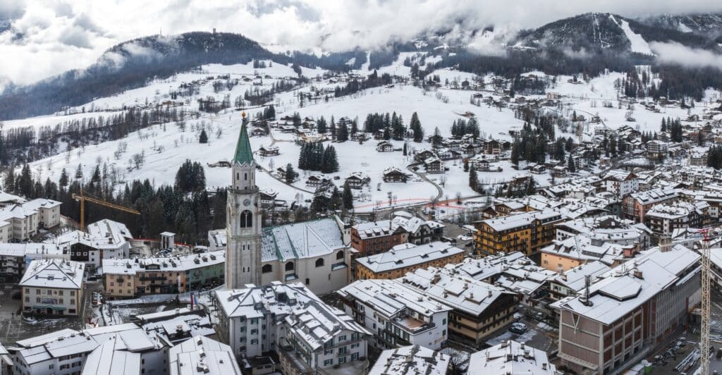 A snowy alpine town with a tall church tower at its center, surrounded by buildings and hillside homes beneath cloudy, snow-covered mountains.
