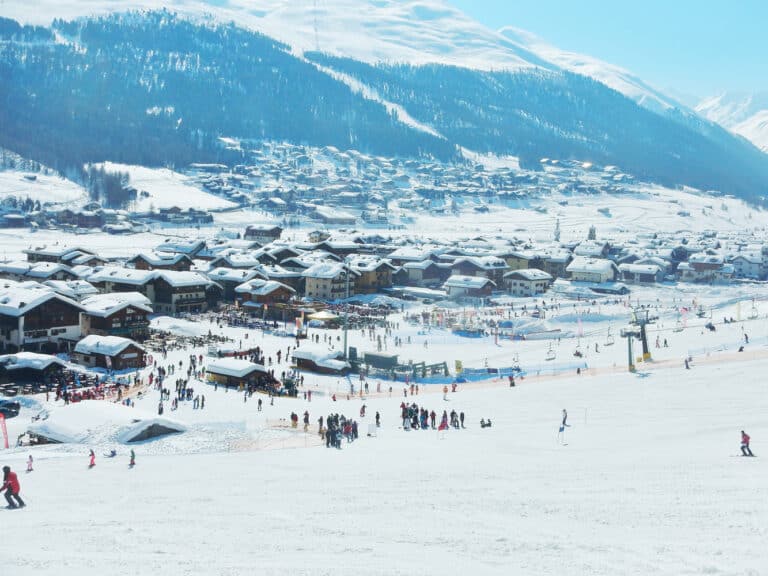 A wide shot of a snow-covered mountain village resort in the Italian Alps with skiers and people gathered at the bottom of the ski slope on a sunny day.
