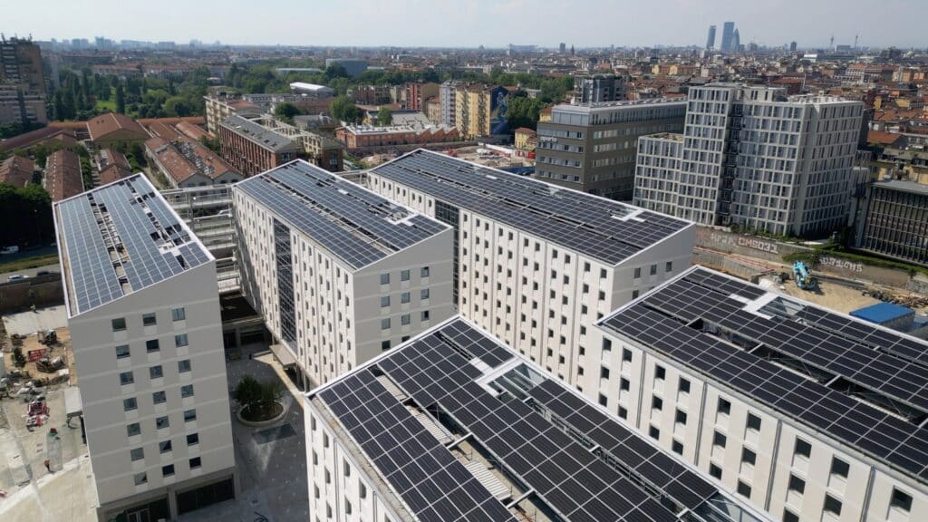 A group of modern residential buildings with large solar panel arrays on their rooftops, set within an urban district overlooking a wide cityscape.