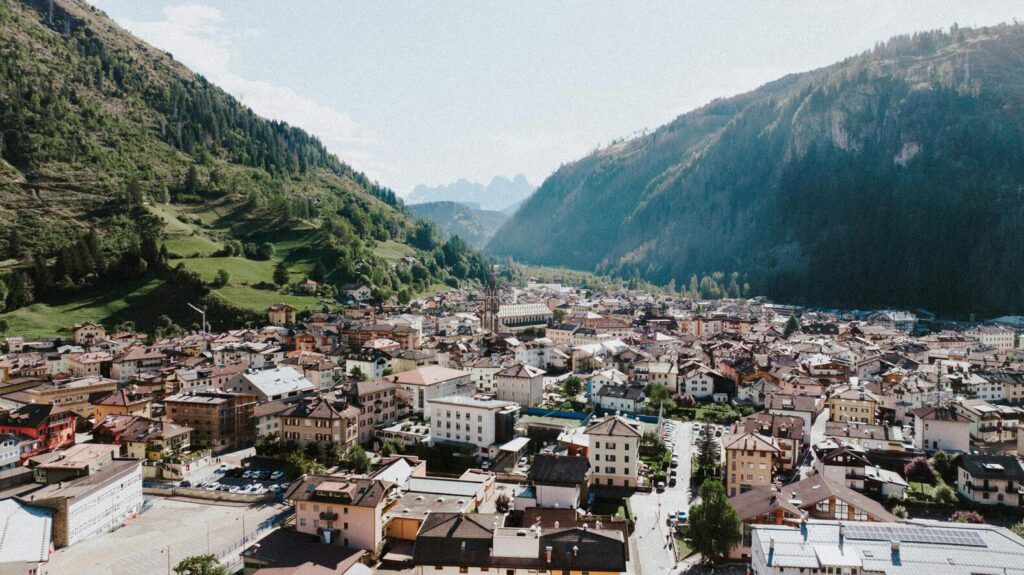 Aerial wide shot of a European mountain town nestled in a valley with steep, green, forested slopes on either side and light haze in the distant peaks.