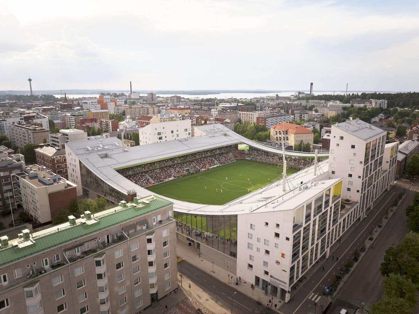 Panoramic aerial shot of a football stadium surrounded by city streets, residential blocks, and waterfront in the distance.