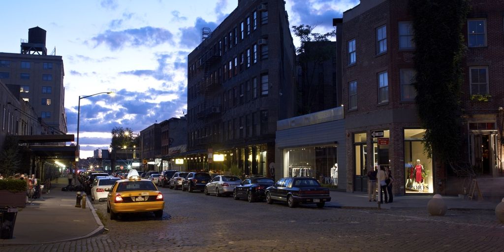 A yellow taxi cab drives down a cobblestone street in New York City at dusk, lined with brick buildings, parked cars, and illuminated storefronts.