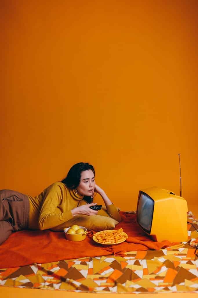 Woman in yellow outfit watching vintage TV with snacks in yellow-themed room.