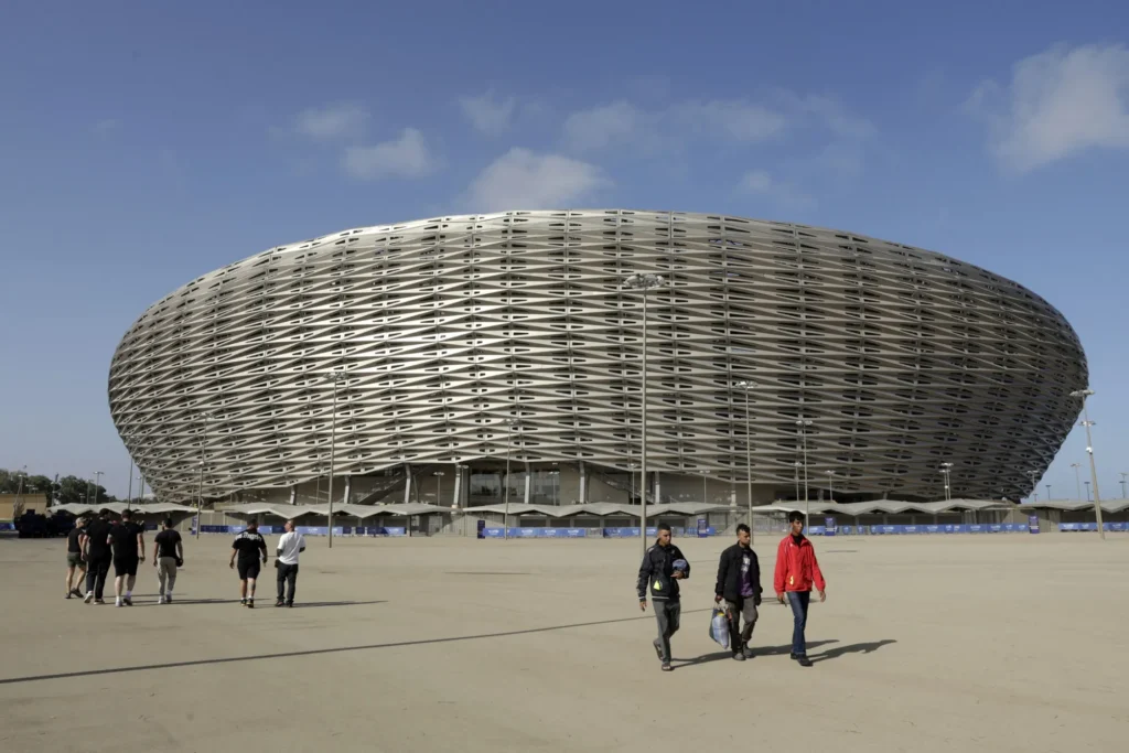 A wide exterior shot of the modern, oval-shaped Baku Olympic Stadium in Azerbaijan, featuring a woven, metallic facade under a blue sky, with groups of people walking on the dusty ground in the foreground.