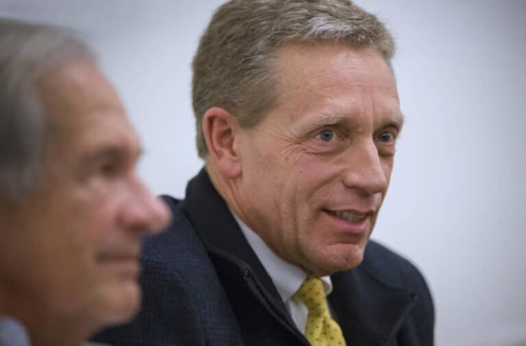Close-up, side profile shot of a middle-aged Caucasian man with graying hair and blue eyes, smiling slightly while wearing a dark jacket and a yellow patterned tie. Another person's out-of-focus face is visible in the foreground.