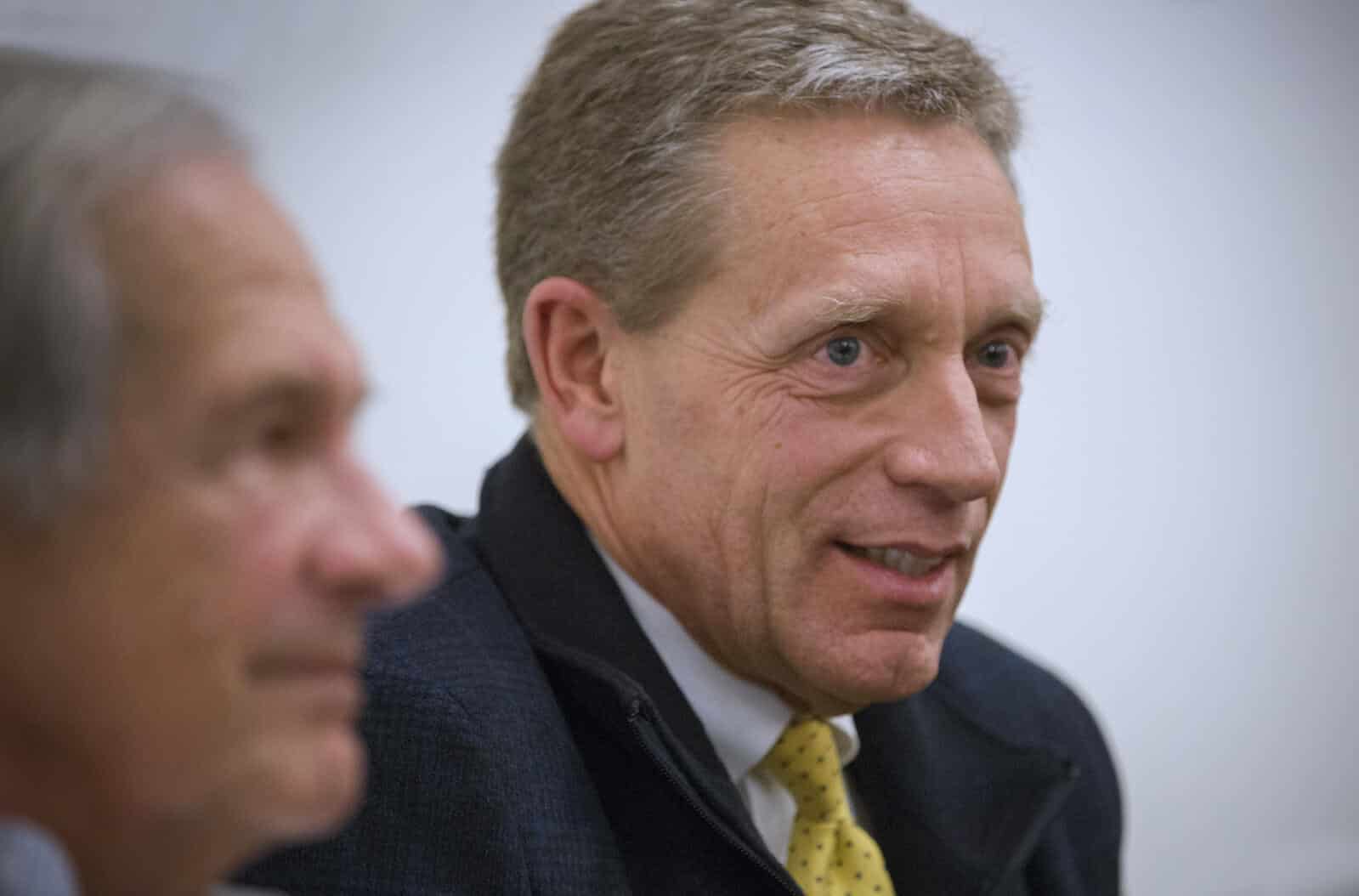 Close-up, side profile shot of a middle-aged Caucasian man with graying hair and blue eyes, smiling slightly while wearing a dark jacket and a yellow patterned tie. Another person's out-of-focus face is visible in the foreground.