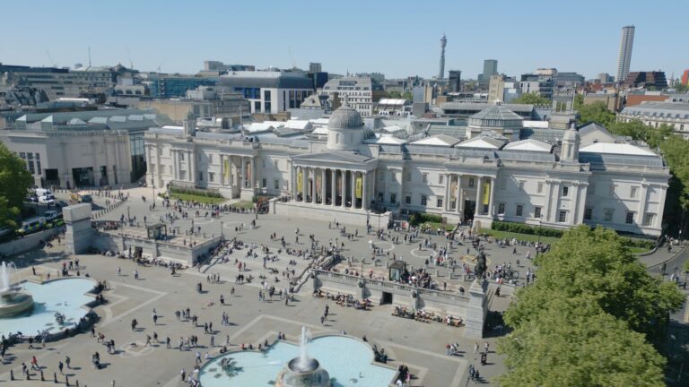 An aerial view of Trafalgar Square in London on a sunny day, showing a large crowd of people. The National Gallery, a grand neoclassical building with columns and a central dome, dominates the center of the square. In the foreground, part of one of the square's fountains is visible. The background shows the city skyline with various modern and historic buildings, including the BT Tower in the distance.