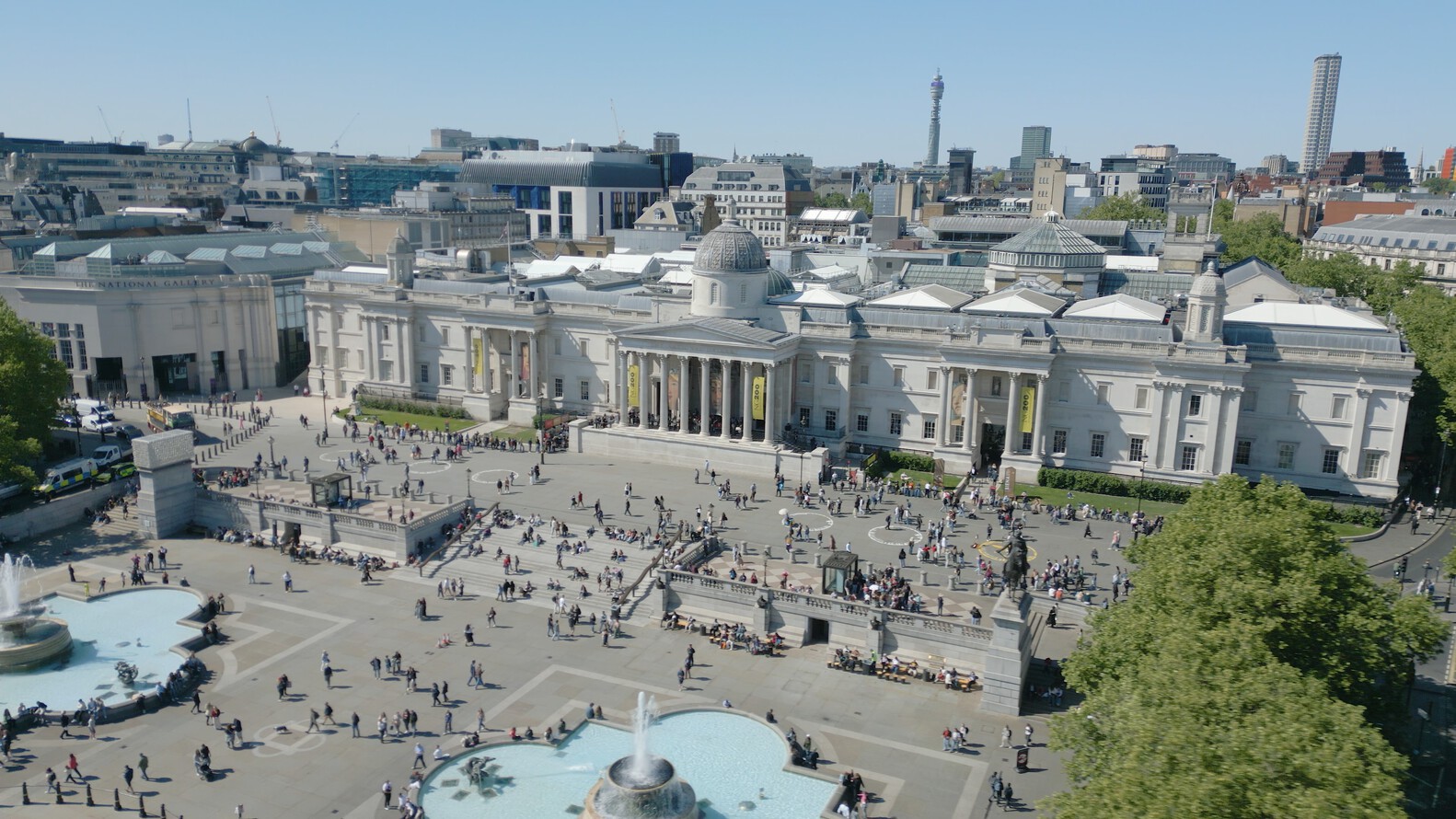 An aerial view of Trafalgar Square in London on a sunny day, showing a large crowd of people. The National Gallery, a grand neoclassical building with columns and a central dome, dominates the center of the square. In the foreground, part of one of the square's fountains is visible. The background shows the city skyline with various modern and historic buildings, including the BT Tower in the distance.