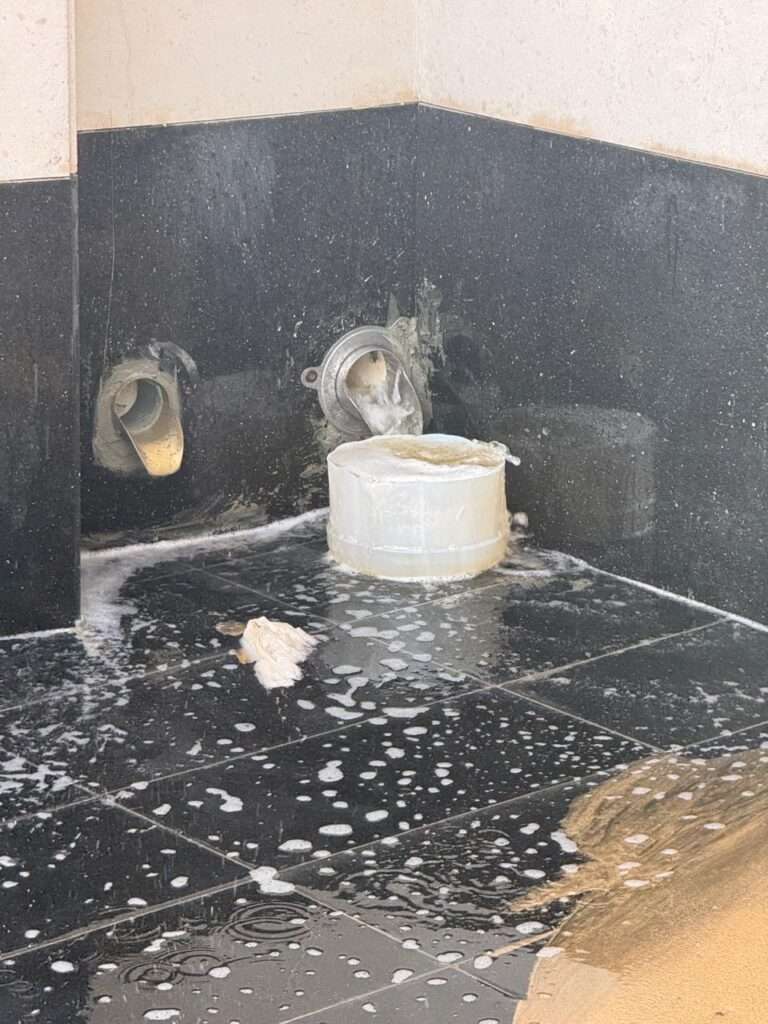 Overflowing rainwater from a wall scupper being caught by a plastic bucket due to drainage system failure.